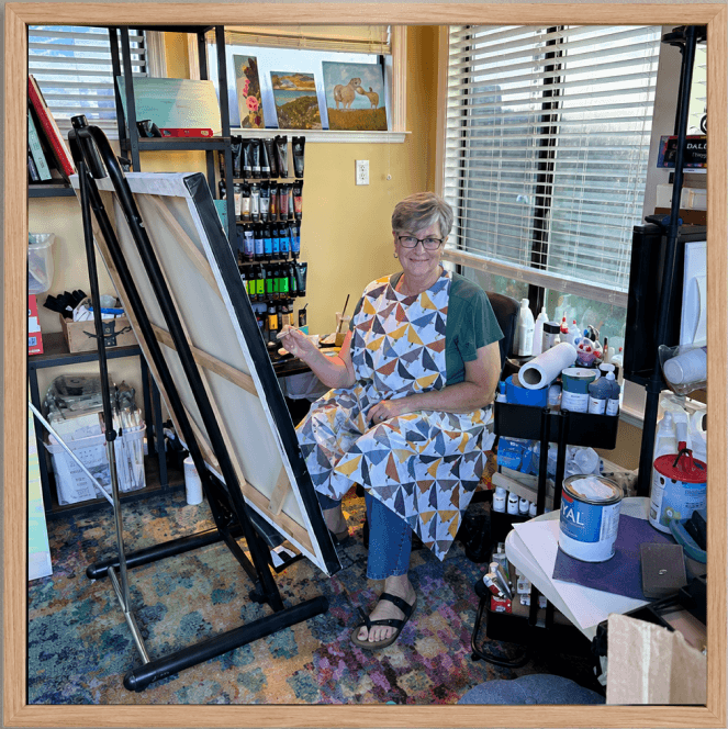 Connie painting at an easel in her studio, surrounded by materials and finished work